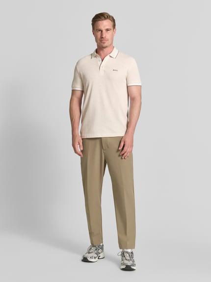 Męski model nosi kremową koszulkę polo Regular Fit Poloshirt aus reiner Baumwolle Modell 'PADDY' von BOSS Green, jasnobeżowe spodnie, szare buty sportowe, tło jednolite jasnoszare, Ganzkörperansicht von vorne.