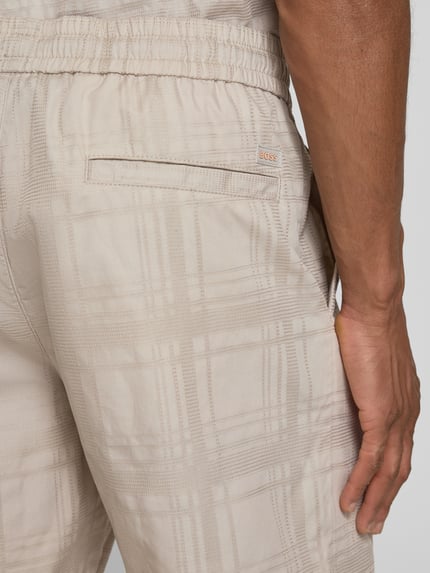 Mężczyzna nosi Tapered Chinoshorts aus reiner Baumwolle Modell 'SANDREW_6_SHORTS' von BOSS Orange, struktura, kolor jasnobeżowy, detal tylnej kieszeni, krój tapered fit, materiał czysta bawełna, zdjęcie z tyłu, zbliżenie na górną część spodenek.