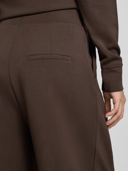 Frauenmodel trägt Barrel Fit Sweatpants mit elastischem Bund von Christian Berg Woman, brauner Material-Mix mit glatter Textur, Nahaufnahme Hüfte. Material-Mix mit Modal-Anteil, Bundfalten und elastischer Bund sichtbar.