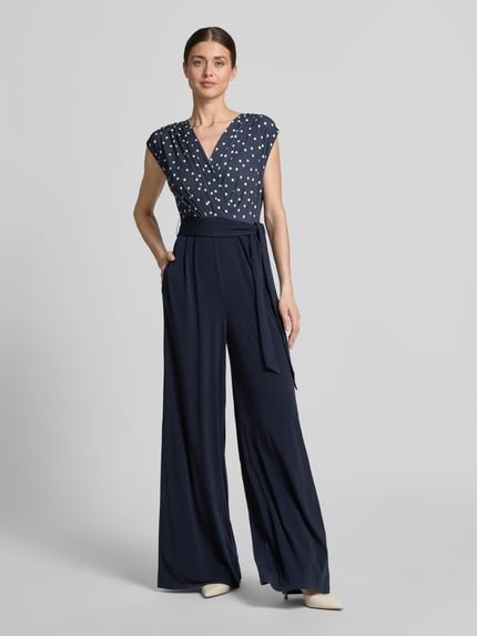 Vrouwenmodel draagt Regular Fit Jumpsuit met V-hals van Vera Mont, donkerblauw met witte stippen, elastische materiaalmix, mouwloos, getailleerde pasvorm, plooien, tailleband, ruglengte maat 36: 39 cm, borstwijdte: 44 cm, mouwlengte: 18 cm, totale lengte: 160 cm, binnenbeenlengte: 93 cm. Ganzlichaamsaanzicht van voren.