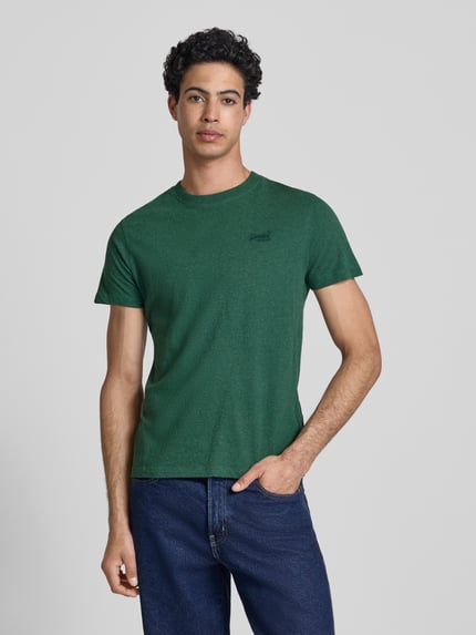 Männermodel trägt Regular Fit T-Shirt mit Logo-Stitching von Superdry in Dunkelgrün, reine Baumwolle, Rundhalsausschnitt, Jeans, Ganzkörperansicht von vorne.