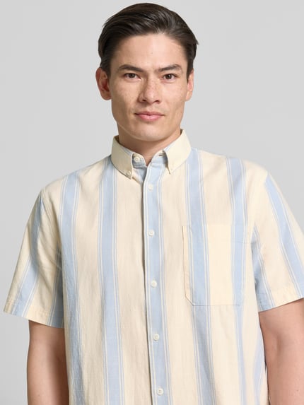 Männermodel trägt Relaxed Fit Freizeithemd aus reiner Baumwolle van Armedangels met blauw-wit gestreept allover-motief, resortkraag, borstzak, korte mouwen, twill textuur, doorknoopsluiting, logo- en labeldetails, pure katoen. Nahaufnahme bovenlichaam.