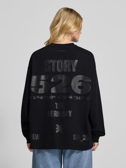 Frauenmodel trägt Oversized Longsleeve mit Logo von Karo Kauer in Schwarz, reine Baumwolle, lockere Passform, Rundhalsausschnitt, Rückenansicht, Rückenlänge S: 72 cm.