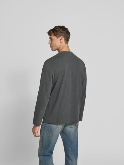 Mężczyzna model nosi Relaxed Fit Longsleeve aus Baumwolle od Forét, kolor szary, efekt znoszenia, wyhaftowane logo, okrągły dekolt, długość tyłu w rozmiarze M: 71 cm, widok całej sylwetki z tyłu.