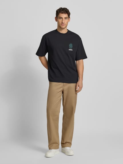 Männermodel trägt schwarzes T-Shirt mit Rundhalsausschnitt Modell 'Gary' von Wood Wood, Relaxed Fit, Label-Print, Ganzkörperansicht von vorne.