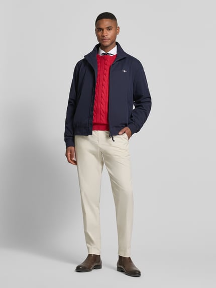 Männermodel trägt Jacke mit Stehkragen und Zweiwege-Reißverschluss von Gant aus Material-Mix mit Baumwoll-Anteil, Regular-Fit, Eingrifftaschen und Label-Stitching, marineblau, kombiniert mit beigen Hosen, Ganzkörperansicht von vorne.