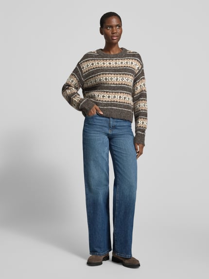 Frauenmodel trägt Pullover mit überschnittenen Schultern Modell 'VMISOLA' von VERO MODA, mehrfarbige Jacquard-Strickoptik, geometrisches Allover-Muster, Cropped-Länge, Rundhalsausschnitt, lange Ärmel, strukturierte Textur, gerippte Abschlüsse, Ganzkörperansicht von vorne.