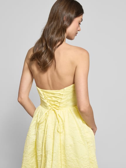 Frauenmodel trägt Minikleid mit floralem Allover-Stitching von Hey Kyla in Pastellgelb, tailliert, schulterfrei, Rückenansicht, Detailaufnahme.