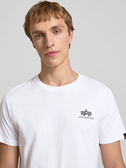 Männermodel trägt T-Shirt mit Logo und Rundhalsausschnitt von Alpha Industries in Weiß, reine Baumwolle, Regular Fit, Nahaufnahme Oberkörper.