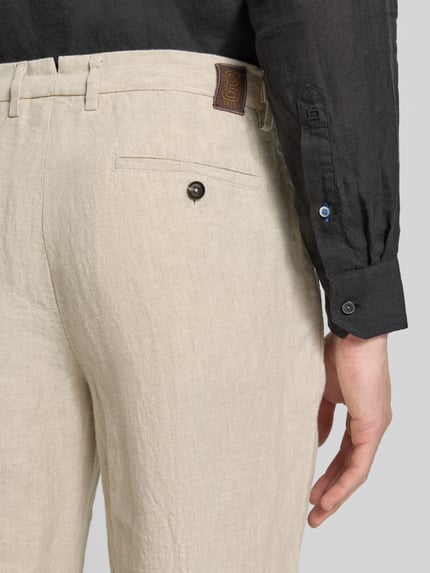 Mężczyzna nosi Leinenhose mit französischen Eingrifftaschen od g1920, len, krój straight fit, jasnobeżowa, wpuszczane kieszenie, długość wewnętrzna nogawki 79 cm, szerokość w rozmiarze 32/32: 44 cm, detal z guzikiem i zapięcie na haftkę i zamek błyskawiczny, widok z tyłu, zbliżenie na kieszenie.