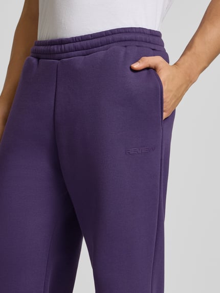Männermodel trägt Loose Fit Sweatpants mit Seitentaschen von Review aus Baumwoll-Mix in Lila, Label-Print, Nahaufnahme Oberkörper.
