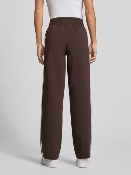 Frauenmodel trägt Regular Fit Sweathose mit Logo-Detail von Adidas Originals in Braun, unifarben, Teilungsnähte, seitliche Taschen, Ganzkörperansicht Rückansicht.