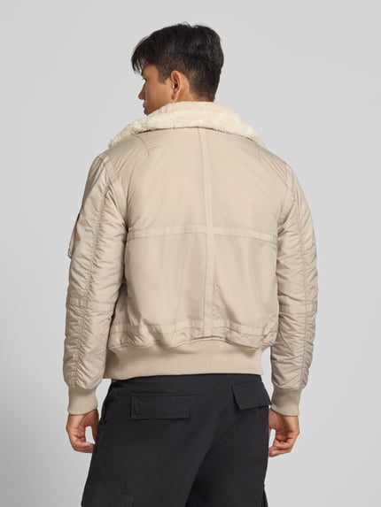 Männermodel trägt Bomberjacke mit Label-Patch und Kentkragen von Alpha Industries in Beige mit Teddyfellkragen, Rückenansicht, Ganzkörperansicht Rückansicht. Regular Fit, Reißverschluss, gerippte Abschlüsse, Reißverschlusstasche am Ärmel, Eingrifftaschen mit Druckknopfverschluss.