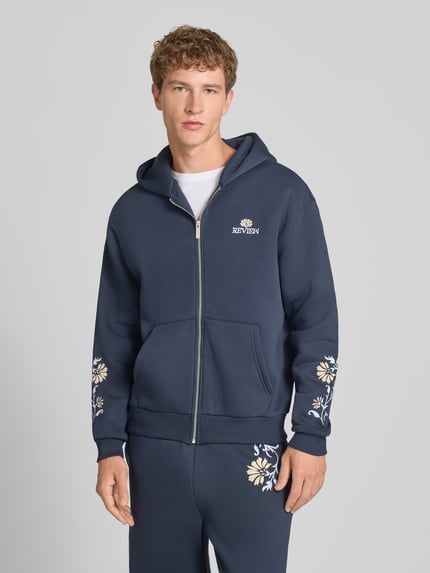 Männermodel trägt Relaxed Fit Sweatjacke mit Kapuze von REVIEW in Dunkelblau, florale Stickerei, Baumwoll-Mix, Ganzkörperansicht von vorne.