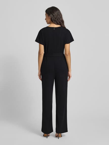 Frauenmodel trägt Jumpsuit mit V-Ausschnitt von Calvin Klein Womenswear, schwarzer Material-Mix mit Stretch-Anteil, lockerer Schnitt, Taillengürtel, Nahtreißverschluss, Ganzkörperansicht Rückansicht.