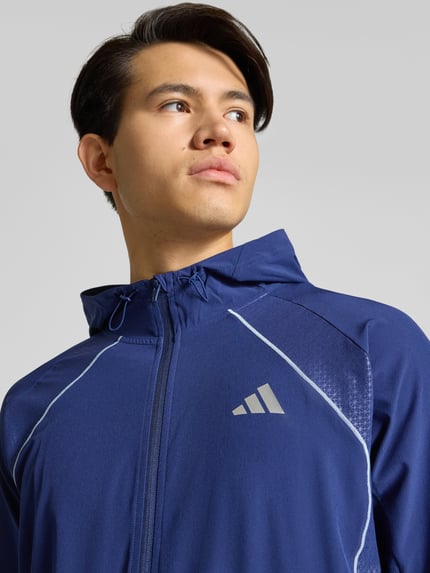 Męski model nosi Regular Fit Kurtka treningowa z nadrukiem z logo Model 'Tech' od adidas Training, elastyczna mieszanka materiałów, kaptur z tunel, wykończenia elastyczne, kieszeń kangurka, zamek błyskawiczny, kolor granatowy, detaliczna faktura, długość tyłu 70 cm, długość rękawa 62 cm, nahaufnahme górnej części ciała.