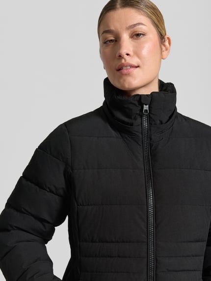 Frauenmodel trägt Jacke mit abnehmbarer Kapuze Modell 'ECLIPSE' von NAUTGÅRD in Schwarz, mit sichtbarem Label-Patch und Zweiwege-Reißverschluss, glatte gesteppte Textur, Nahaufnahme Oberkörper, gerader Schnitt, gestrickte Ärmelabschlüsse, seitliche Eingrifftaschen mit Druckknopfverschluss.