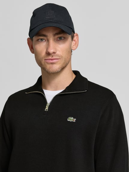 Männermodel trägt Regular Fit Troyer aus reiner Baumwolle von Lacoste in Schwarz mit Reißverschluss und Krokodil-Logo, Nahaufnahme Oberkörper. Herren Troyer von Lacoste.