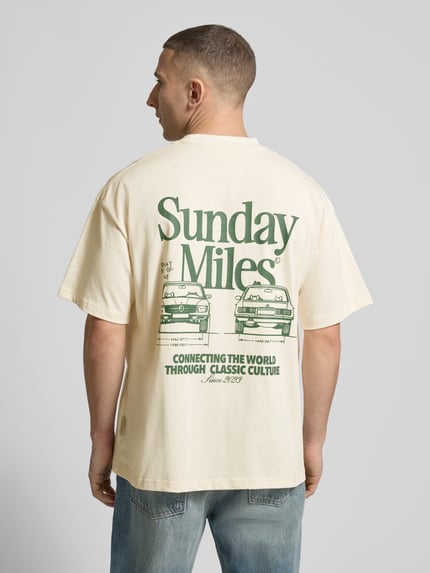 Männermodel trägt T-Shirt aus Baumwolle Modell 'Sunday Miles' von ANOTHER COTTON LAB in Creme mit grünem Print, Rückenansicht, Ganzkörperansicht Rückansicht.