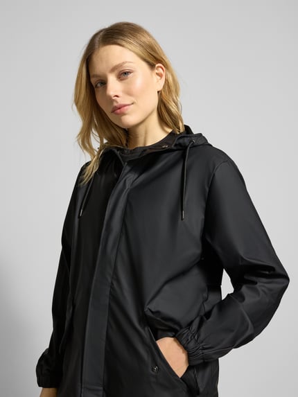 Frauenmodel trägt Regular Fit Parker mit Kapuze Modell 'Fishtail' von Rains in Schwarz, glatte Textur, Pattentaschen, elastische Ärmelabschlüsse, Ganzkörperansicht von vorne. Rückenlänge bei Größe S: 104 cm, Schulterbreite: 45 cm, Brustweite: 55 cm, Ärmellänge: 65 cm.