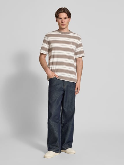 Mannenmodel draagt Regular Fit T-shirt uit katoenmix van Calvin Klein Jeans met all-over motief, gestreept patroon, logo-stitching detail, ronde hals, korte mouw, regular fit pasvorm, kleur beige-wit-bruin Ganzkörperansicht von vorne.