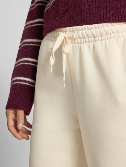 Kobieta nosi Wide Fit Sweatpants aus Baumwoll-Mix Modell 'Iconic' od Gina Tricot, elastyczny pas ze sznurkiem, kremowy kolor, miękka bawełniana faktura, widoczny fragment dłoni, kontekst: damskie spodnie dresowe z bawełnianego miksu, szeroki krój, długość nogawki w rozm. S: 80 cm, zdjęcie zbliżenie na górną część spodni.