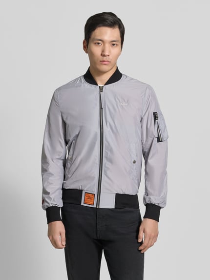 Männermodel trägt silbergraue Bomberjacke mit Label-Patch und Eingrifftaschen von Original Bombers, Stoff leicht glänzend, Ganzkörperansicht von vorne, Gerader-Schnitt, Reißverschluss, Reißverschlusstasche am Ärmel, Druckknopfverschluss, Gerippte-Abschlüsse, Rückenlänge bei Größe M: 67 cm, Schulterbreite: 43 cm, Brustweite: 53 cm, Ärmellänge: 68 cm.