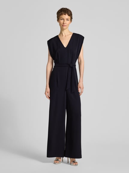 Frauenmodel trägt Jumpsuit mit V-Ausschnitt und Bindegürtel von comma, dunkle Farbe, glatte Textur, Regular Fit, Ganzkörperansicht von vorne.