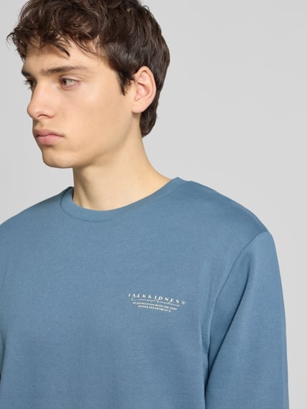 Männermodel trägt Sweatshirt mit Label-Print von Jack & Jones in Blau, Rundhalsausschnitt, lockerer Schnitt, Baumwoll-Mix, Nahaufnahme Oberkörper.
