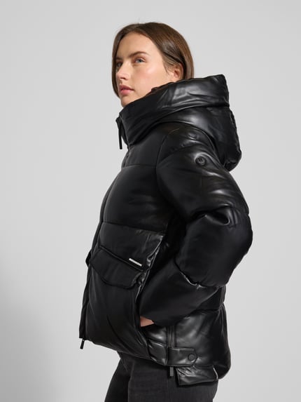 Frauenmodel trägt schwarze khujo Steppjacke mit Kapuze Modell 'AURORA', Pattentaschen, Reißverschluss, Label-Detail, seitliche Ansicht