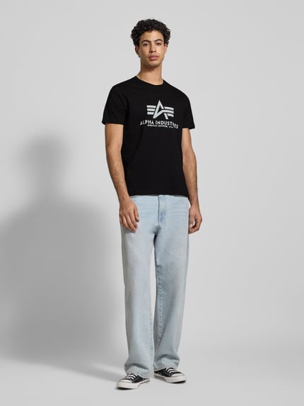 Männermodel trägt T-Shirt mit Label-Print von Alpha Industries, Reine Baumwolle, Gerader Schnitt, Rundhalsausschnitt, Ganzkörperansicht von vorne.