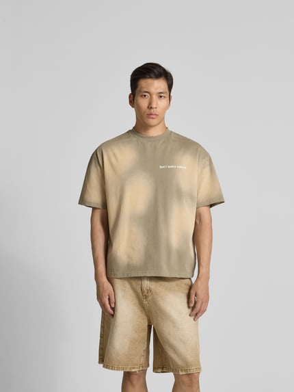 Męski model nosi T-Shirt mit Label-Print od Don't Waste Culture z bawełny, prosty krój, okrągły dekolt, nadruk z logo, całkowita długość tyłu 65 cm przy rozmiarze M, kolor beżowy z nieregularnymi plamami, krótkie rękawy, widok całej sylwetki z przodu.