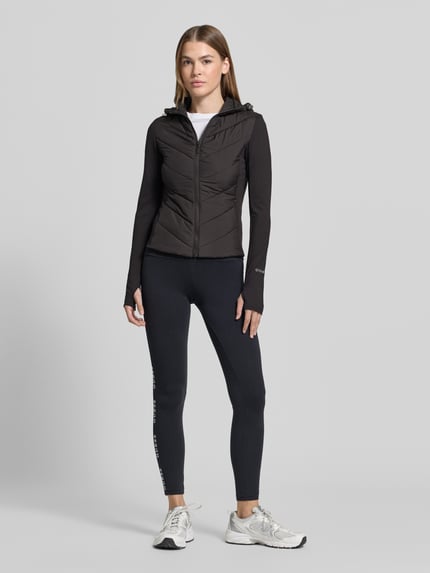 Modelka nosi czarną Steppjacke mit Kapuze Modell 'GRAZIA' von GUESS Activewear, krój slim fit, gładka tekstura, widok całej sylwetki z przodu, długość pleców w rozmiarze S 55 cm, szerokość ramion w rozmiarze S 38 cm, szerokość w rozmiarze S 40 cm, długość rękawa w rozmiarze S 68 cm.