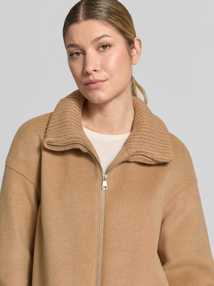 Frauenmodel trägt Mantel aus Woll-Mix mit Reißverschlusstaschen von White Label in Camel, grobe Strickstruktur am Stehkragen, Nahaufnahme Oberkörper. Regular Fit, glatte Oberfläche, sichtbarer Reißverschluss, Kontext: Rückenlänge 86 cm, Schulterbreite 55 cm, Brustweite 55 cm, Ärmellänge 56 cm.