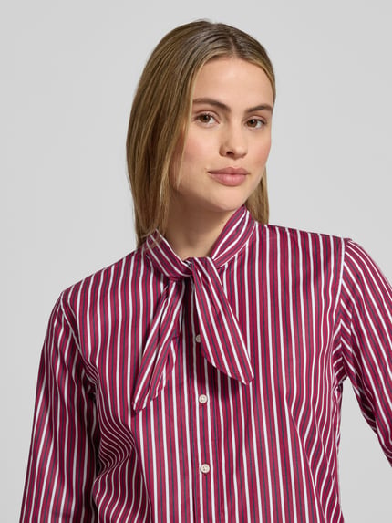 Frauenmodel trägt Hemdbluse mit Knopfleiste Modell 'NUSILA' von Lauren Ralph Lauren, Streifenmuster in Pink und Weiß, Schleifen-Detail, Reine Baumwolle, Lockerer-Schnitt, Nahaufnahme Oberkörper.