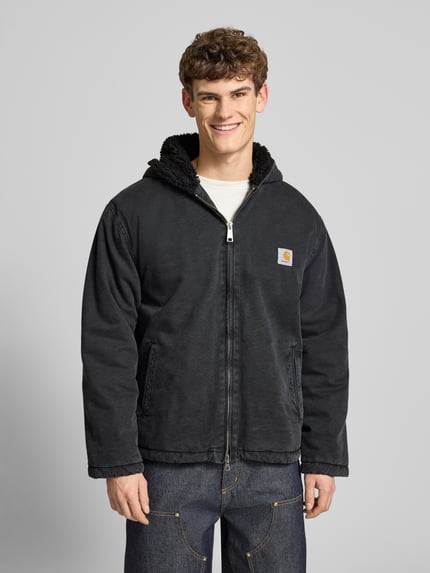 Männermodel trägt Jacke mit Zweiwege-Reißverschluss Modell 'MITCH' von Carhartt Work In Progress aus Baumwolle, Regular Fit, Kapuze, Eingrifftaschen, dunkles einfarbiges Design, Ganzkörperansicht von vorne.