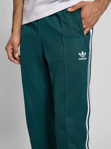 Männermodel trägt Sweatpants mit elastischem Bund van adidas Originals in donkergroen met witte strepen, katoenmix, label-stitching, ritszakken, achterzak; detailaufnahme heup en bovenbeen.