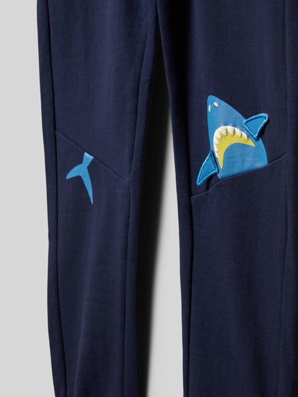 Produktfoto, Regular Fit Sweatpants mit Motiv-Print von Blue Seven in Blau, Reine Baumwolle, Eingrifftaschen, elastischer Bund mit Tunnelzug, elastische Beinabschlüsse, Innenbeinlänge 53 cm bei Größe 116, weiche Textur, sichtbar Hai-Motiv-Print auf der Tasche.