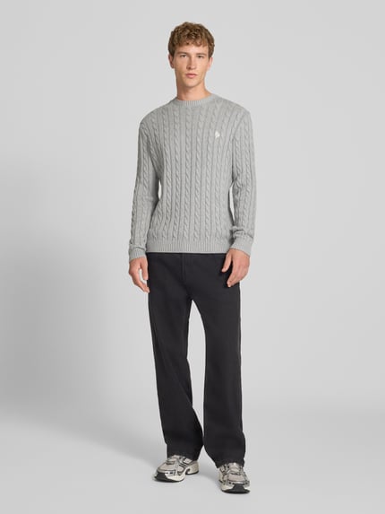 Męski model nosi Slim Fit Strickpullover reine Baumwolle od REVIEW, szary, ścieg warkoczowy, wyhaftowane logo, ściągacze, krój slim fit, bawełna, długość tyłu 73 cm, długość rękawów 70 cm, widok całej sylwetki z przodu.