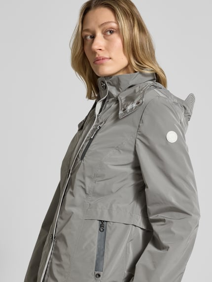 Kobiece model prezentuje Jacke mit Kapuze und Eingrifftaschen von Gil Bret, krój regular fit, kaptur, listwa guzikowa, kieszenie zapinane na zamek błyskawiczny, weather protection, długość tyłu w rozmiarze 36: 67 cm, szerokość ramion w rozmiarze 36: 40 cm, szerokość klatki piersiowej w rozmiarze 36: 46 cm, długość rękawów: 64 cm, odcień szarości, gładka faktura, zbliżenie na górną część ciała, widok z przodu.