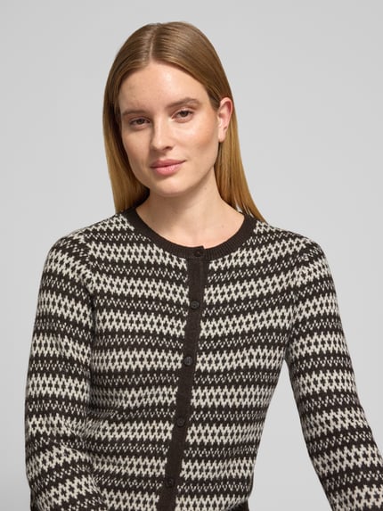 Frauenmodel trägt Cardigan mit Zierknöpfen von VERO MODA, geometrisches Allover-Muster in Schwarz und Creme, Strickoptik, Nahaufnahme Oberkörper.