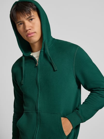 Männermodel trägt Regular Fit sweatjack van katoenmix met labeldetail van Superdry in donkergroen, kangoeroezak, capuchon met tunnelkoord, elastische boorden, ruglengte maat M 65 cm, mouwlengte maat M 65 cm, Nahaufnahme Oberkörper.