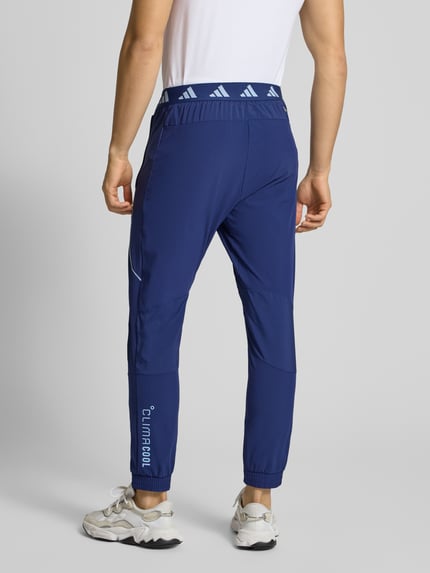 Mannelijk model draagt Slim Fit Sweatpants met Logo-Print Model 'Tech' van adidas Training in blauw, elastische materiaalmix, logoprint, slim fit, elastische band en enkelboorden, binnenbeenlengte maat M 75 cm, ganzkörperansicht van achteren.