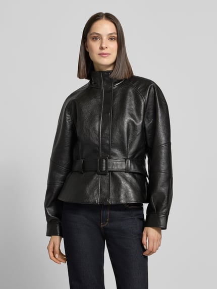 Frauenmodel trägt Jacke in Leder-Optik mit Gürtel Modell 'Jaclyn' von Neo Noir, Schwarz, strukturierte Oberfläche, Ganzkörperansicht von vorne. Relaxed Fit, Stehkragen, Abmessungen Größe 36: Rückenlänge 57 cm, Schulterbreite 41 cm, Brustweite 51 cm, Ärmellänge 62 cm.