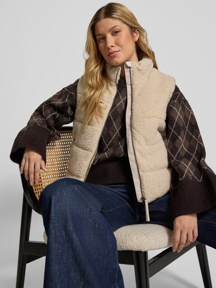 Modelka prezentuje Steppweste aus Teddyfell mit Zweiwege-Reißverschluss od camel active w kolorze kremowym, krój regular fit, detaliczny widok górnej części ciała z przodu, długość tyłu 55 cm, szerokość ramion 40 cm, szerokość w rozmiarze 36: 52 cm, stójka, kieszenie na zamek.