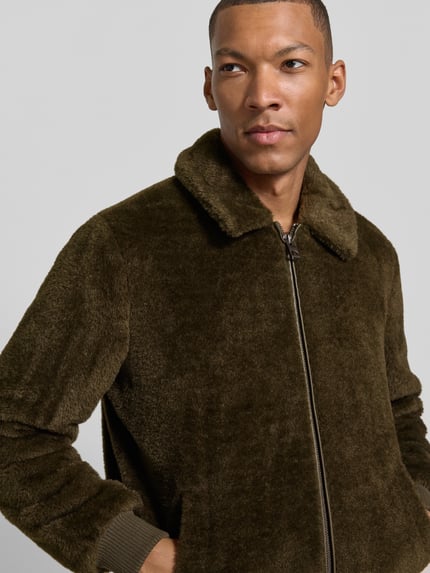 Męski model nosi Regular Fit Bomberjacke mit Zweiwege-Reißverschluss od GUESS, kolor oliwkowy, Kołnierzyk typu kent, prążkowane wykończenia, wpuszczane kieszenie, Długość tyłu w rozmiarze M: 66 cm, Szerokość ramion w rozmiarze M: 43 cm, Szerokość w rozmiarze M: 57 cm, Długość rękawów: 61 cm, Ganzkörperansicht z przodu