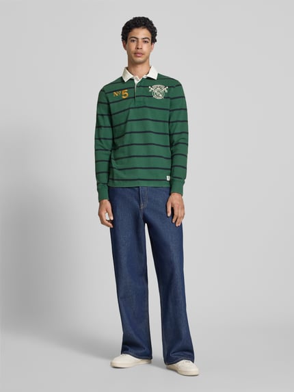 Männermodel trägt Regular Fit Rugby Shirt mit Logo- und Motiv-Stitching von Superdry in Grün mit Streifen und Label-Patch, Reine Baumwolle, Polokragen, kurze Knopfleiste, Rückenlänge bei Größe M: 70 cm, Ganzkörperansicht von vorne.