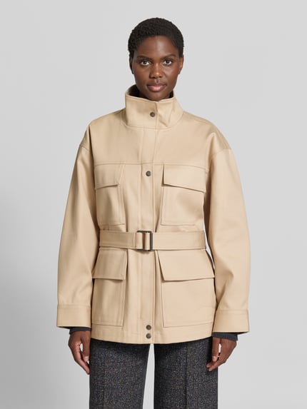 Vrouwenmodel draagt Trenchcoat met tailleriem van Anine Bing in beige gewaxte katoen, getailleerde pasvorm, vier klepzakken, doorlopende drukknoopsluiting, maat S: ruglengte 74 cm, borstwijdte 56 cm, mouwlengte 58 cm, Ganzekörperaanzicht van voren.