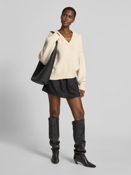 Vrouwenmodel draagt Oversized Pullover in tricotlook van black palms, katoenmix, oversized fit, V-hals, elastische boorden, ruglengte maat S 64 cm, zandkleur, gladde en gebreide texturen, Ganzkörperansicht von vorne.