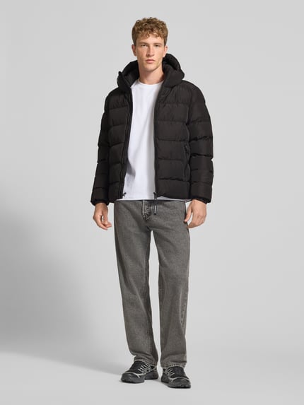 Männermodel trägt Steppjacke mit Label-Stitching und Kapuze von Superdry in Schwarz, kombiniert mit Jeans und Sneakers, Ganzkörperansicht von vorne. Herren Steppjacke von Superdry mit geradem Schnitt, Kapuze, Kordelzug, Reißverschluss, Seitentaschen, elastischen Ärmelabschlüssen.